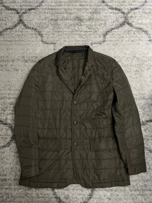 Olivgrön quiltad trenchcoat - Stilren olivgrön quiltad trenchcoat med tre knappar framtill och två fickor med lock. Jackan har klassisk krage och lätt vadderad design som ger en modern look. Perfekt för dig som vill ha en trendig och funktionell jacka med snygg passform.