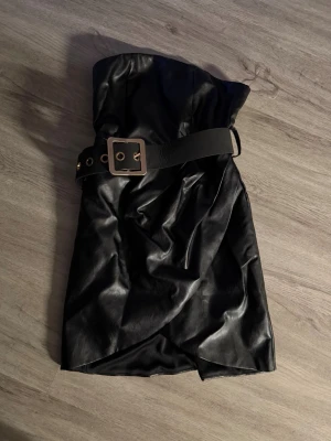 Svart axelbandslös klänning med bälte - Snygg svart axelbandslös klänning i skinnimitation med bredt bälte och stor guldfärgad spänne. Kort omlottmodell som ger en cool och edgy look. Perfekt för dig som vill sticka ut och gillar statement-detaljer.
