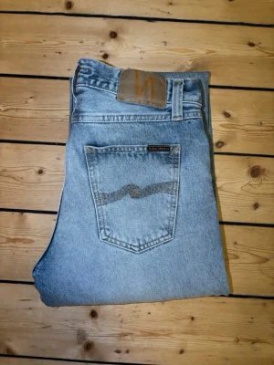 Ljusblå raka jeans från Nudie - Säljer ett par ljusblå jeans från Nudie med klassisk rak passform och snygga detaljer på bakfickorna. Jeansen har en patch i brunt läder baktill och är tillverkade i slitstark denim. Perfekta för en avslappnad och trendig look.