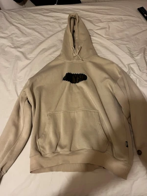 Beige hoodie från distict fortysix - Snygg beige hoodie från district fortysix med brun broderad logga på bröstet. Klassisk känguruficka framtill och justerbar huva med dragsko. Tillverkad i mjukt bomullsmaterial som är skönt mot huden. Perfekt för en avslappnad streetwear-look.