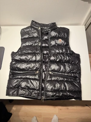 Moncler Väst - Moncler Gui Gilet Väst, finns ett litet hål, se bild 2.           Sätt en svart liten tejpbit över det så är det inget som syns på håll. Dragkedjan funkar inte, därav säljs denna billigt till någon som kanske är på en tajt budget🙂Jag är 178cm lång och den passar perfekt.
