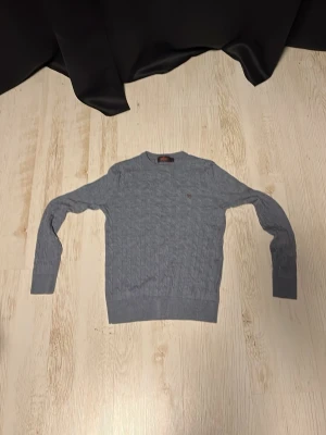  kabelstickad tröja  - Snygg grå kabelstickad tröja från Morris med rund halsringning och diskret broderad logga på bröstet. Tröjan har ribbade muddar vid ärmslut och nederkant. Perfekt för dig som gillar klassisk och stilren design.