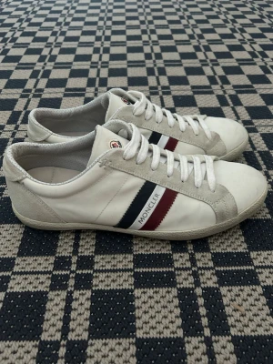 Vita Moncler sneakers med ränder - Snygga vita sneakers från Moncler med klassiska röda och marinblå ränder på sidan samt Moncler-logga på plösen. Skorna har lågt skaft, vita snören och detaljer i mocka runt tån och hälen. Perfekta för dig som gillar stilrena och exklusiva sneakers.