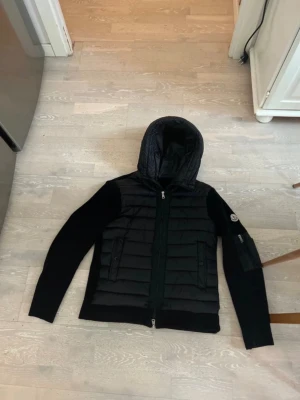 Svart pufferjacka från Moncler - Snygg svart pufferjacka från Moncler med quiltad front och huva. Jackan har släta ärmar, två dragkedjor fram och en Moncler-logga på ärmen. Materialet är en mix av dun och textil, vilket ger både värme och stil. Perfekt för kalla vinterdagar. Funkar både S och M. Pris kan diskuteras!
