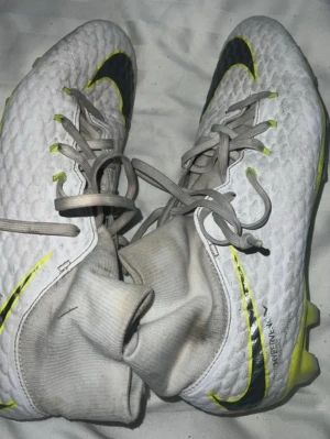 Nike Hypervenom vita fotbollsskor - Säljer ett par vita Nike Hypervenom fotbollsskor med högt skaft och snörning. Skorna har gula och svarta detaljer, samt mönstrad ovandel i syntetmaterial. Sulan har gula dobbar för bra grepp på planen. Perfekta för dig som vill sticka ut på matchen!