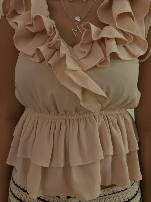Beige volangblus med omlottdetalj - Söt beige blus med volanger och omlottdesign framtill. Toppen är ärmlös och har resår i midjan för en snygg siluett. Tillverkad i ett lätt och luftigt material som ger en romantisk vibe.