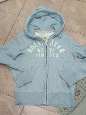 Ljusblå hoodie från Hollister - Ljusblå zip-hoodie från Hollister med vit text 'Hollister Wave Finals' på bröstet. Hoodien har huva, två fickor fram och ribbade muddar. Mjuk insida och dragkedja hela vägen. Perfekt för en avslappnad och chill stil.