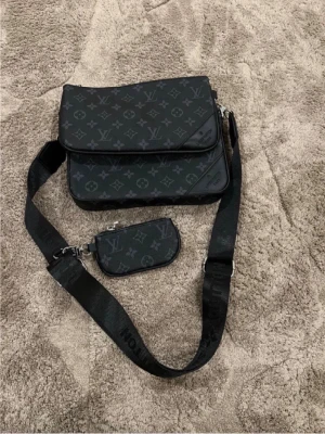 Louis Vuitton svart axelväska monogram - Snygg svart axelväska från Louis Vuitton med klassiskt monogrammönster i grått. Väskan har justerbar axelrem och en mindre avtagbar ficka. Tillverkad i slitstarkt canvasmaterial med detaljer i svart. Perfekt för dig som vill ha en trendig och praktisk väska.