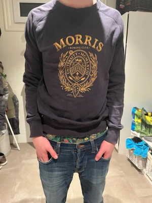 Morris mörkblå sweatshirt med tryck - Mörkblå sweatshirt från Morris med stort guldigt universitetsinspirerat tryck på bröstet. Tröjan har rund hals, ribbade muddar och avslappnad passform. Perfekt för dig som gillar klassisk college-stil och snygga detaljer.