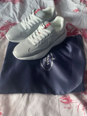 Vita sneakers från Prada - Snygga vita sneakers från Prada med klassisk design och röda detaljer på plösen. Skorna har snörning och är tillverkade i en mix av skinn och syntetmaterial för en clean och modern look. Perfekta för dig som vill ha stilrena och exklusiva sneakers.