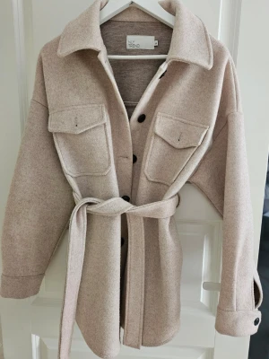 Beige cardiganjacka från NLY Trend - Stilren beige cardiganjacka från NLY Trend med knytskärp i midjan, stora bröstfickor och svarta knappar. Jackan har klassisk krage och är tillverkad i ett mjukt, ull-liknande material. Perfekt för lager på lager och trendiga outfits.Stor i storlek
