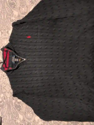 Mörkblå kabelstickad tröja Polo Ralph Lauren - Kabelstickad mörkblå tröja från Polo Ralph Lauren med halv dragkedja och röd broderad logga på bröstet. Tröjan har ribbade muddar vid ärmslut och nederkant samt hög krage. Klassisk och tidlös design i mjukt material. Strl M men passar bra på S