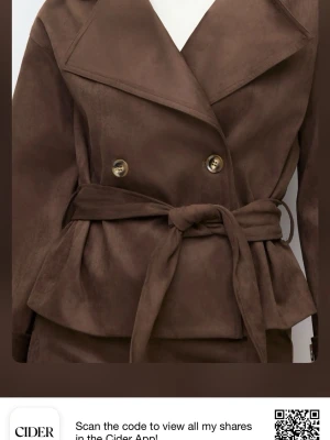 Brun trenchcoat med knytskärp från Cider - Stilren brun trenchcoat från Cider i mjukt material med bred krage, dubbelknäppning och knytskärp i midjan. Jackan har dekorativa knappar på ärmsluten och en figurnära passform som ger en snygg siluett. Perfekt för dig som vill ha en trendig och klassisk look.