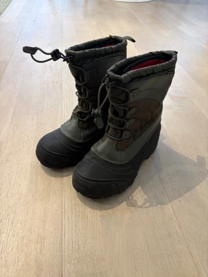 Svarta vinterstövlar från The North Face - Tuffa svarta vinterstövlar från The North Face med högt skaft, grov sula och snörning framtill. Utsidan är i slitstark textil och gummi, insidan har röd vaddering för extra värme. Perfekta för kalla dagar och snöiga äventyr.