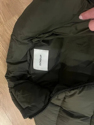 Mörkgrön dunjacka från DONDUP - Snygg mörkgrön dunjacka från DONDUP med dragkedja och justerbar dragsko nedtill. Jackan har en klassisk puffer-modell och är tillverkad i ett glansigt material som ger en modern look. Perfekt för kalla vinterdagar.