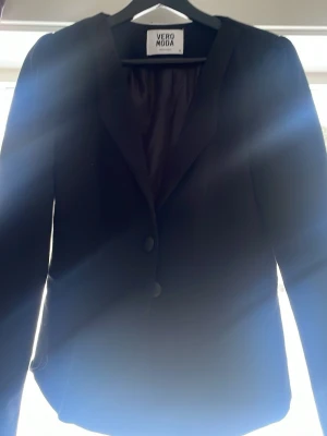 Svart kavaj från Vero Moda - Stilren svart kavaj från Vero Moda med klassisk krage och två knappar framtill. Figurnära passform och helfodrad insida. Perfekt att matcha med skjorta eller t-shirt för en snygg look.
