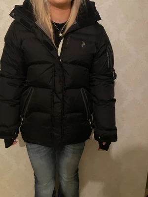 Svart dunjacka från Peak Performance - Säljer en svart dunjacka från Peak Performance med huva och dragkedja framtill. Jackan har två snedställda fickor med dragkedja och en extra ficka på ärmen. Perfekt för kalla vinterdagar och riktigt skön att bära. Storlek M