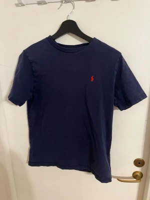 Mörkblå t-shirt från Ralph Lauren - Klassisk mörkblå t-shirt från Ralph Lauren med röd broderad logga på bröstet. T-shirten har rund halsringning och korta ärmar. Tillverkad i bomull för en skön känsla och enkel stil.