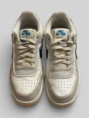 Nike Air Force 1 vit/grå sneakers - Klassiska Nike Air Force 1 sneakers i vitt och beige med blå detaljer. Skorna har lågt skaft, perforerad tåbox och ikonisk Swoosh-logga på sidan. Ovandelen är i läder och textil, med vadderad plös och bekväm innersula. Perfekta för en streetwear-look.