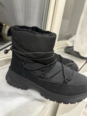 Svarta moon boots från Bench - Säljer ett par svarta moon boots från Bench med tjock sula och snörning runt hela skon. Ovandelen är i vadderat material och insidan ser riktigt mjuk ut. Perfekta för kalla dagar när du vill hålla stilen och värmen. Endast använda 2 gånger 