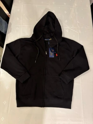 Svart zip hoodie från Polo Ralph Lauren - Klassisk svart hoodie med dragkedja från Polo Ralph Lauren. Har huva med dragsko, ribbade muddar och fickor framtill. Liten röd logga broderad på bröstet. Tillverkad i mjuk bomullsblandning för skön känsla.