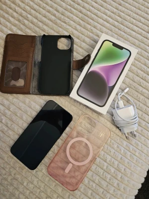 iPhone med tillbehör och skal - Säljer en svart iPhone från Apple med slimmad design och glasbaksida. Medföljer originalkartong, laddare med USB-C kabel, ett brunt plånboksfodral i läder, ett svart skal och ett genomskinligt MagSafe-kompatibelt skal i plast. Perfekt för dig som vill ha både skydd och stil.