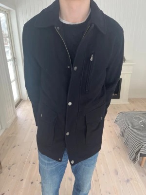 Navy ullrock med fickor och dragkedja - Stilren navy ullrock från kända märket Brooks brothers.  Klassisk krage, tryckknappar och dragkedja. Rocken har två stora framfickor med lock och en bröstficka med dragkedja. Quiltat innerfoder för extra värme. Jackor från märket ligger på ca 4000-6000kr