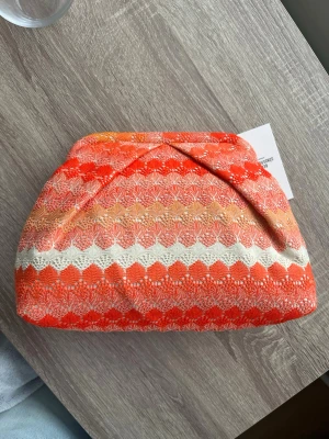 Mönstrad orange clutch - Säljer en unik clutch i tyg med vågmönster i orange, rött, beige och vitt. Väskan har en mjuk, vadderad form och ett snyggt vikt lock. Perfekt för dig som vill sticka ut med färg och mönster.
