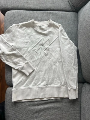 Vit sweatshirt med broderad logga - Vit sweatshirt i mjukt material med ribbade muddar vid ärmslut och nederkant. Framsidan har en broderad logga i vitt som ger en clean och stilren look. Rund halsringning och relaxed fit gör den enkel att matcha till olika outfits.