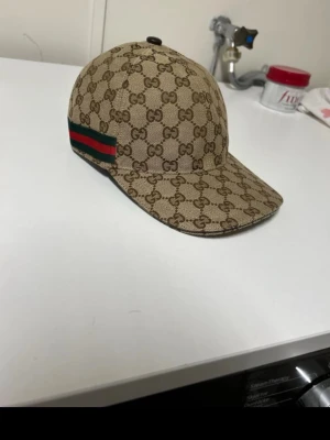 Gucci beige keps med GG-mönster - Säljer en beige keps från Gucci med klassiskt GG-monogram över hela kepsen. På sidan finns den ikoniska gröna och röda randen. Justerbar rem bak i svart läder och böjd skärm. Tillverkad i canvasmaterial som ger en exklusiv känsla. Säljs billigt och den är typ oanvänt har använt den max ett par gånger då och då inget mer därför säljs den för bra pris vid snabb affär går priset att diskuteras