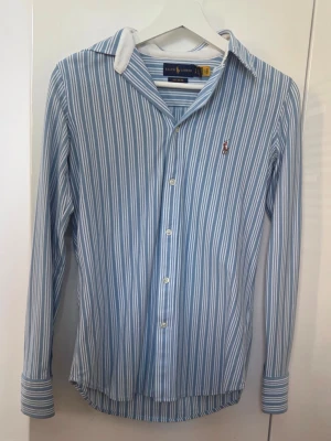 Blåvit randig skjorta från Ralph Lauren - Snygg blå och vit randig skjorta från Ralph Lauren med klassisk krage och knappar framtill. Skjortan har lång ärm och ikoniska broderade loggan på bröstet. Tillverkad i mjuk bomull som känns skön mot huden.