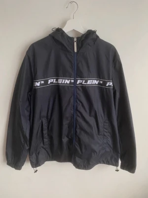 Svart vindjacka från Plein - Snygg svart vindjacka från Plein med huva och dragkedja framtill. Jackan har ett vitt band med Plein-logga tvärs över bröstet och två fickor med dragkedja. Tillverkad i lätt och vindtätt material, perfekt för blåsiga dagar.Philipp Plein jacka Storlek L Använd 2-3 gånger Köpt för : 7555kr  Pris går att diskutera Kan skicka mer bilder om det skulle viljas och tveka inte att höra av dig vid frågor☺️