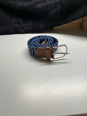 Randigt blått flätat bälte - Snyggt flätat bälte i marinblått och ljusblått med bruna skinn-detaljer och silverfärgat spänne. Perfekt accessoar för att lyfta din outfit med en cool och modern touch.