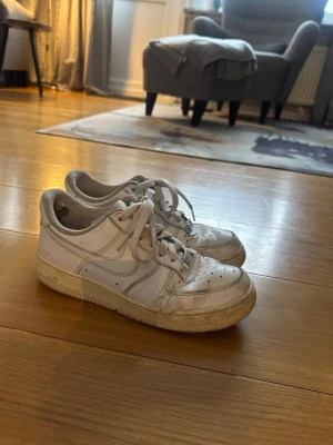 Vita Nike Air Force 1 sneakers - Klassiska vita Nike Air Force 1 sneakers med lågt skaft och perforerade detaljer på tån. Skorna har en robust sula och är tillverkade i läder med textilfoder. Swoosh-logga på sidan och snörning framtill för en tidlös streetwear-look.