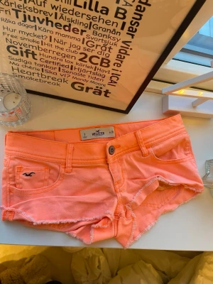 Korta rosa/orange shorts från Hollister - Säljer ett par korta rosa/neon färgade shorts från Hollister (California) kanter och klassisk fickdesign Shortsen har låg midja och är tillverkade i mjuk bomull, perfekta för varma sommardagar. Snygga detaljer med broderad logga på fickan i storlek w26🤍 midjemåttet är 40cm från ena sidan till andra inte runt hela🥰❣️💗