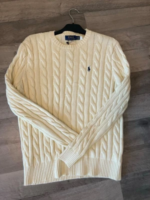Kabelstickad offwhite tröja Polo Ralph Lauren - Kabelstickad tröja i offwhite från Polo Ralph Lauren. Klassisk modell med rund halsringning, ribbade muddar och det ikoniska blå broderade logot på bröstet. Perfekt för dig som gillar tidlös och stilren design.