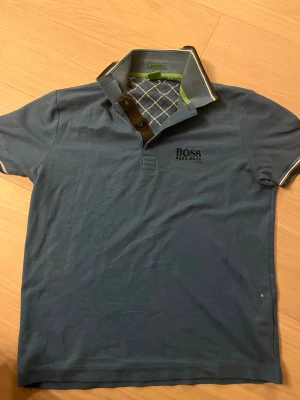 Blå pikétröja från Hugo Boss - Snygg blå pikétröja från Hugo Boss med broderad logga på bröstet. Kragen har randiga detaljer och insidan har ett rutigt mönster i blått och vitt samt en grön kant. Tröjan har korta ärmar och knappar vid halsen.