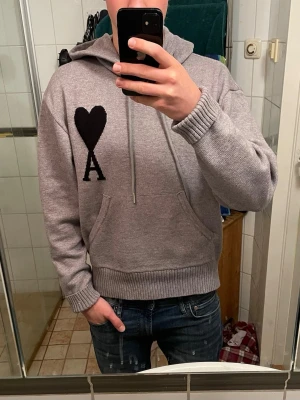 Grå hoodie från Ami Paris - Säljer en grå hoodie från Ami Paris med svart hjärta och bokstaven A på bröstet. Hoodien har huva med dragsko, magficka och ribbade muddar vid ärmslut och nederkant. Tillverkad i mjukt stickat material som känns riktigt skönt.            Priset kan diskuteras såklart!                                         Först till kvarn!                                                                   Kom gärna med prisförslag!!