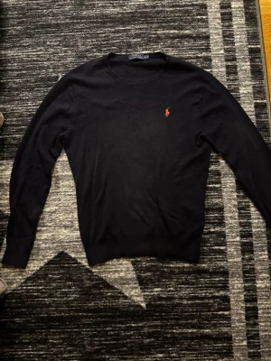Mörkblå tröja Polo Ralph Lauren - Snygg mörkblå stickad tröja från Polo Ralph Lauren med klassisk rund halsringning och den ikoniska röda logotypen broderad på bröstet. Tröjan har långa ärmar och ribbade muddar vid ärmslut och nederkant. Perfekt för en stilren och avslappnad look.