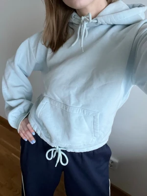 Ljusblå hoodie med dragsko - Säljer en ljusblå hoodie med huva och dragsko både i midjan och vid halsen. Hoodien har en stor magficka och ribbade muddar vid ärmsluten. Perfekt för en avslappnad look. 