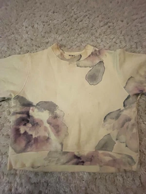 Beige sweatshirt med mönster från Acne - En beige cropped hoodie med längre ärmar från Acne med blommigt mönster. Några få fläckar därav det låga priset.