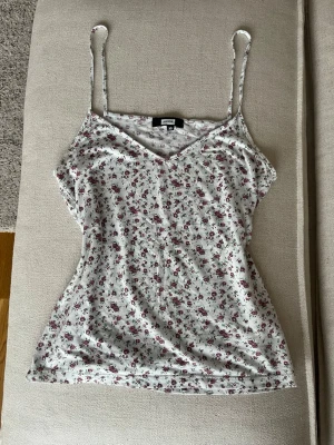 Vit blommig linne från Bik Bok - Söt vit linnetopp från Bik Bok med små röda blommor. Toppen har smala axelband och v-ringning, perfekt för varma dagar. Tunn och mjuk i materialet, lätt att matcha till jeans eller kjol.