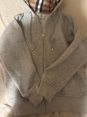 Grå zip hoodie från Burberry 165 cm - Snygg grå zip hoodie från Burberry med klassiskt rutigt foder i huvan. Hoodien har dragsko i huvan, ribbade muddar och fickor framtill. Tillverkad i mjukt bomullsmaterial som känns riktigt skönt mot huden. Perfekt för en avslappnad och trendig look.