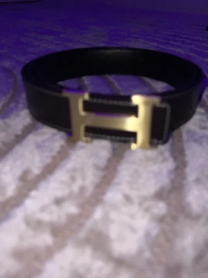 Hërmẽš  bälte - Snyggt brunt skärp  med ikoniskt H-format spänne i guld. Skärpet har vita sömmar längs kanterna och är tillverkat i slätt skinn. Perfekt accessoar för att lyfta vilken outfit som helst.