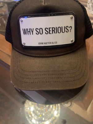 Svart keps John Hatter & Co - Snygg svart truckerkeps från John Hatter & Co med vit metallskylt framtill där det står 'WHY SO SERIOUS?'. Kepsen har mesh på sidorna och bak för extra ventilation och justerbar passform. Perfekt för dig som vill sticka ut med en cool detalj.