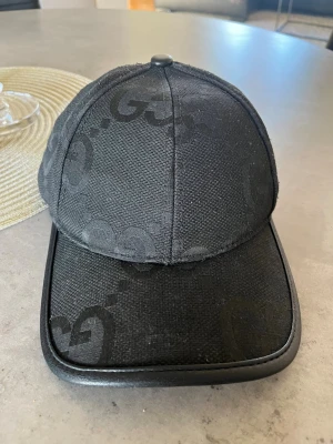 Svart keps från Gucci med GG-mönster - Snygg svart keps från Gucci med det klassiska GG-monogrammet ton-i-ton över hela kepsen. Kepsen har böjd skärm med svart kant i läderlook och justerbart band bak. Tillverkad i Italien och har en exklusiv känsla som passar perfekt till streetwear.