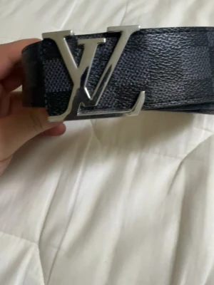  rutigt svart bälte - Snyggt svart bälte från med klassiskt rutigt mönster och stor logga i silver på spännet. Bältet är gjort i skinn och har en exklusiv känsla som lyfter vilken outfit som helst.