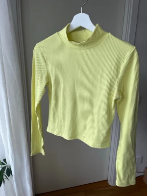 Ljusgul/grön ribbad topp Gina Tricot  - Trendig ljusgul/grön topp med lång ärm och ribbad struktur. Hög hals ger en snygg siluett och den stretchiga bomullsblandningen gör den bekväm att bära. Perfekt att styla med jeans eller kjol för en fräsch look. 
