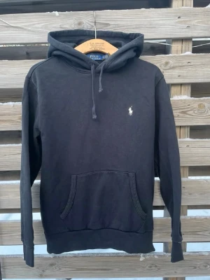 Svart hoodie från Polo Ralph Lauren - Klassisk svart hoodie från Polo Ralph Lauren med vit broderad logga på bröstet. Hoodien har huva med dragsko, stor magficka och ribbade muddar vid ärmar och nederkant. Tillverkad i mjukt bomullsmaterial för en skön och avslappnad look.
