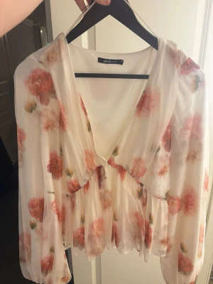Blommig chiffongblus från Gina Tricot - Romantisk blus i tunn, vit chiffong med stora rosa och röda blommor. Djup v-ringning, volangdetaljer och lös passform. Långa, puffiga ärmar med resår vid handlederna ger en luftig och trendig look. Perfekt för att lyfta vilken outfit som helst.